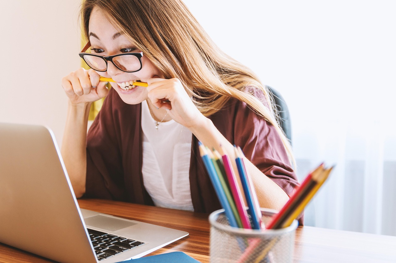 Woman wearing glasses sat at laptop with pencil in her mouth and pencil pot on the desk